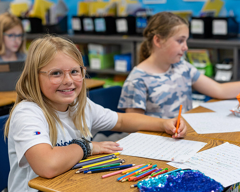 2 Pine Ridge female students working with colored pencils at their desks, one smiling at the camera