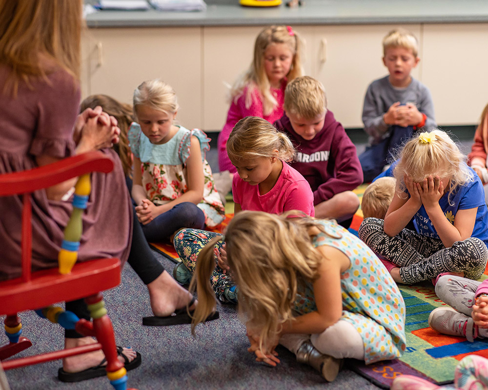 kindergartners praying together with their teacher sitting in a red rocking chair