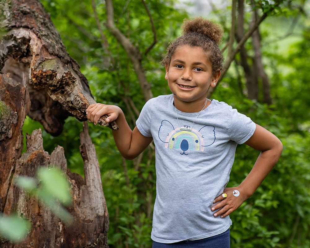 Forest School 1st grader smiling at the camera, hand on hip, holding on to a tree stump