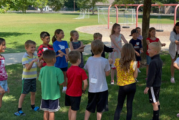 Spanish Immersion elementary camp students playing a game standing in a circle outside with their teacher demonstrating something with her arms.