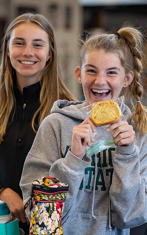 2 middle school girls at lunch smiling, holding up a snack