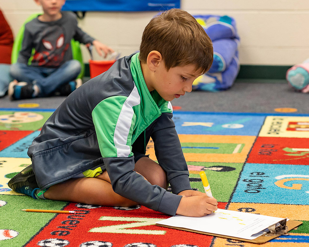 Spanish Immersion kindergarten boy writing on a clipboard on the colorful alphabet rug