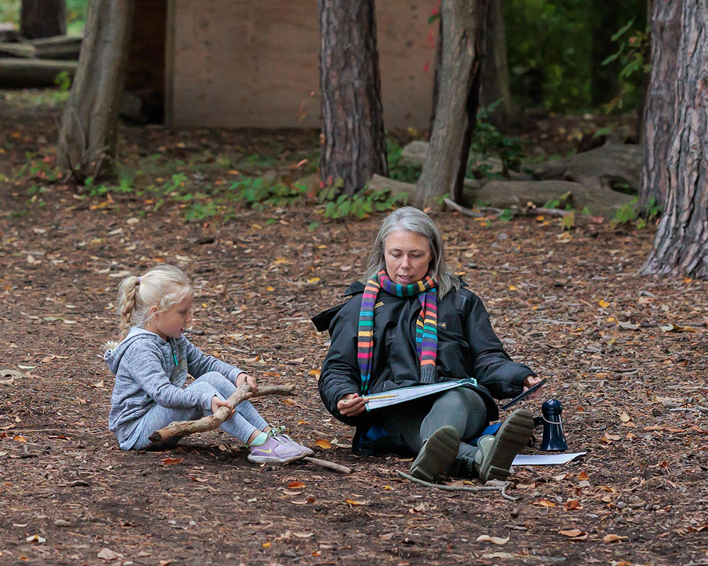 Teacher aide and girl kindergarten student reading together in the forest, sitting on the ground.