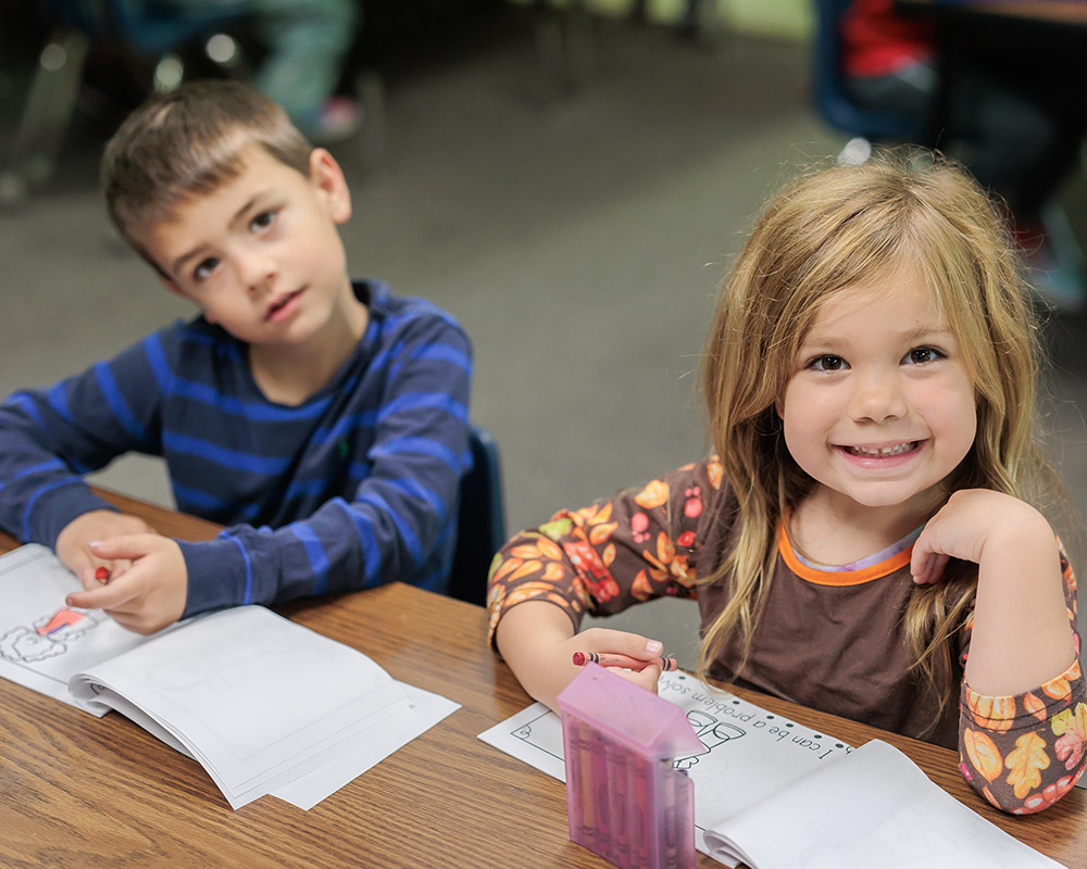 Boy and girl kindergarten students coloring a page, with the girl smiling at the camera