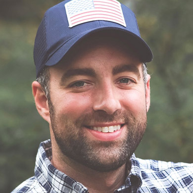 Brad Sawdon smiling at the camera, wearing blue plaid shirt and American flag baseball hat.