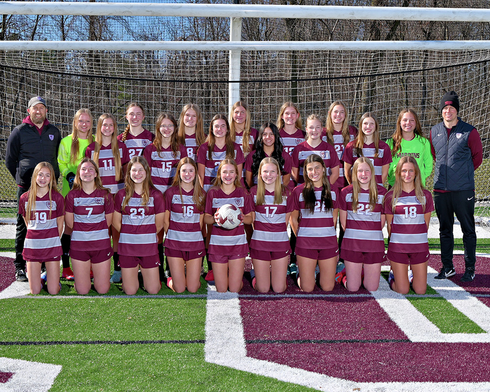 JV girls soccer team standing in front of a soccer goal and smiling at the camera, all in maroon and gray striped uniforms.