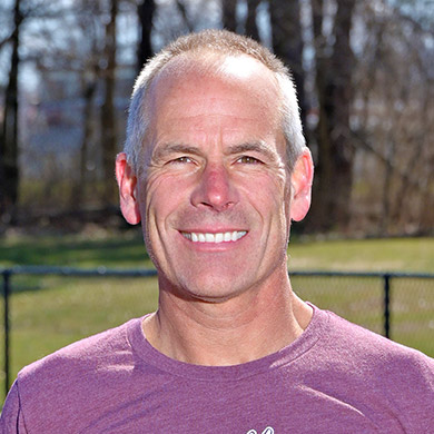Track coach smiling at the camera, while standing on the track in a maroon heathered track t-shirt