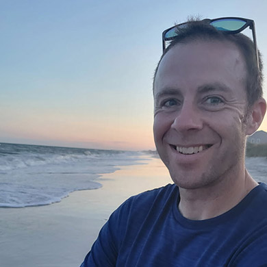 Joe Brunson smiling at the camera with the beach and a sunset in the background.