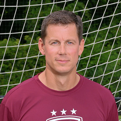 Coach DeBoer standing in front of a soccer goal, looking at the camera