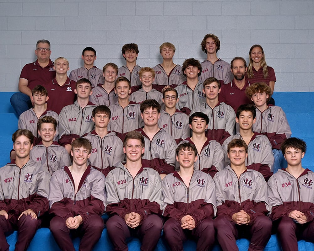 Boys aquatics team sitting in bleachers, smiling at the camera.