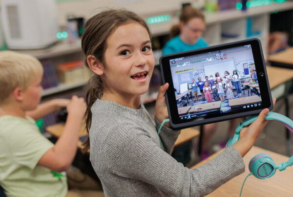 Smiling elementary girl holding up her ipad with photo of students on it.