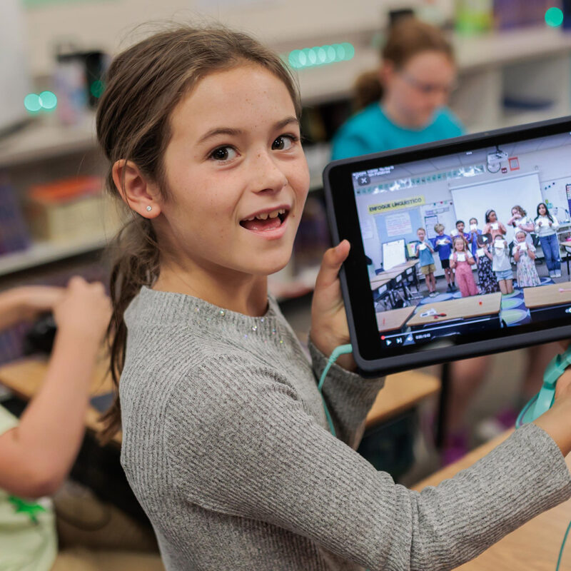 Smiling elementary girl holding up her ipad with photo of students on it.