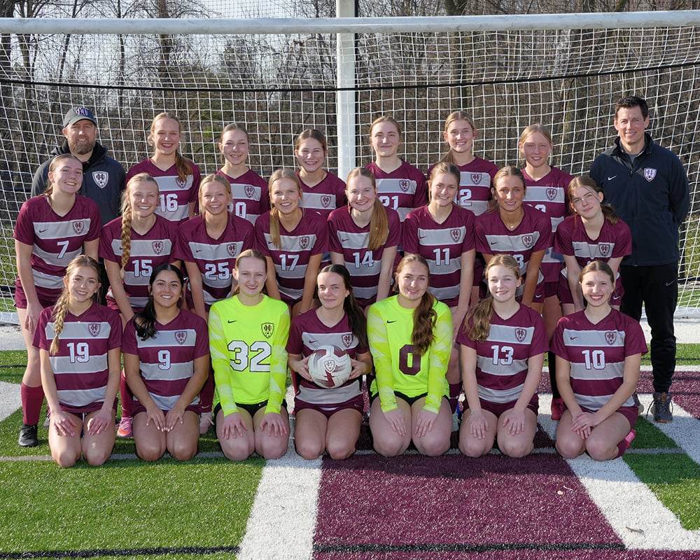 JV girls soccer team standing in front of a soccer goal and smiling at the camera, all in maroon and gray striped uniforms.