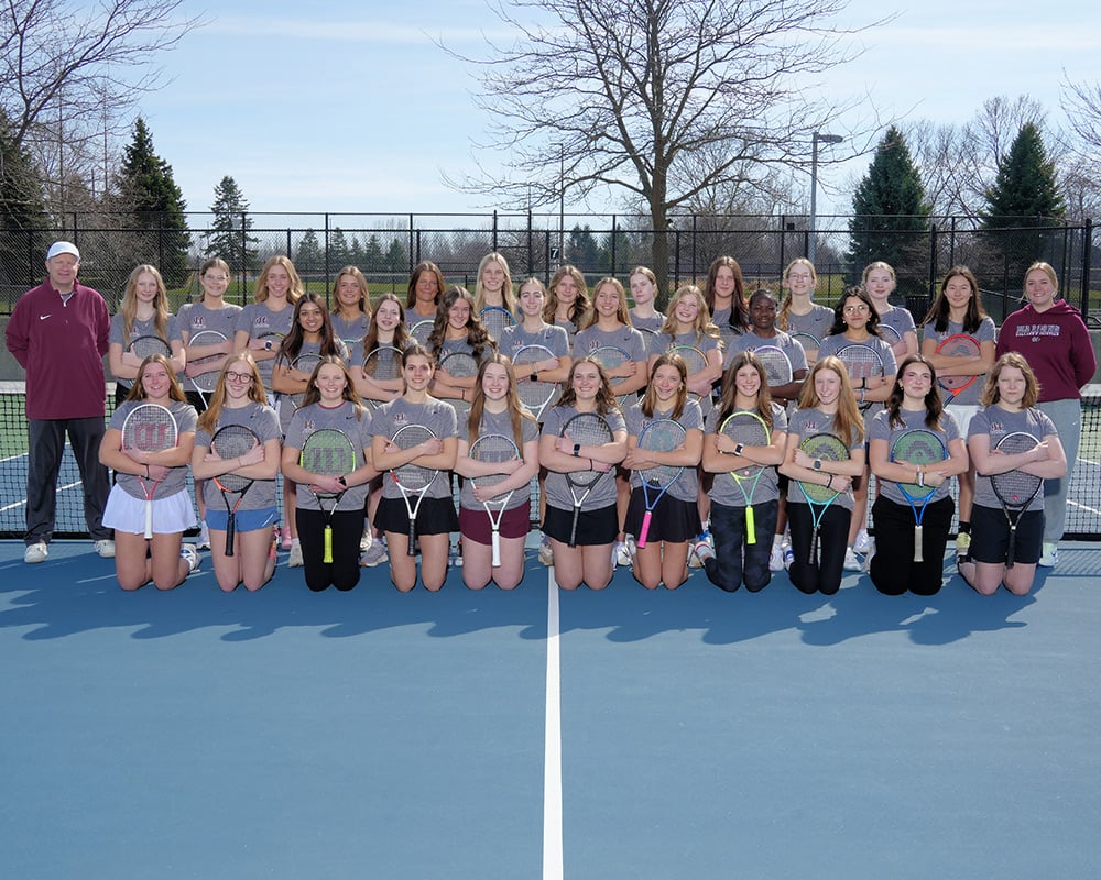 Girls JV tennis team wearing gray uniform tops, while holding their rackets in front of them and smiling at the camera