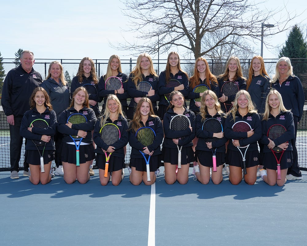 Girls varsity tennis team wearing black uniform dresses and holding their tennis rackets in front of them, while smiling at the camera