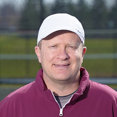 Girls JV tennis coach standing on the tennis courts and smiling at the camera.