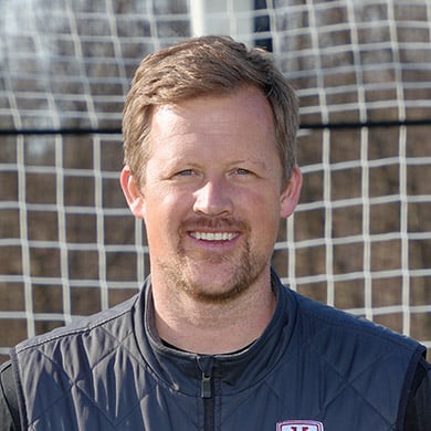 Girls varsity soccer coach standing in front of the soccer goal and smiling at the camera