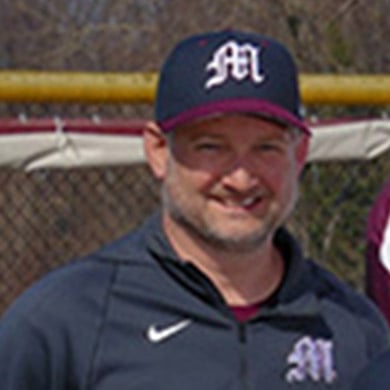 Boys varsity baseball coach smiling at the camera, outside on a sunny day.